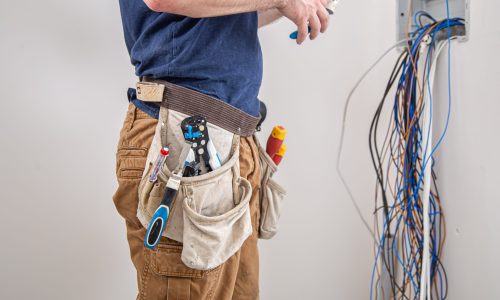 Electrician builder at work, examines the cable connection in the electrical line in the fuselage of an industrial switchboard. Professional in overalls with an electrician's tool.